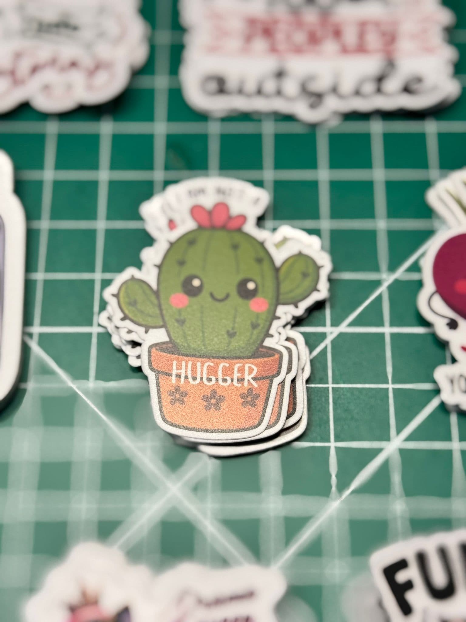 A cartoon cactus with a smiling face and arms, sitting in a pot labeled "HUGGER," is surrounded by various other colorful stickers on a green cutting mat.