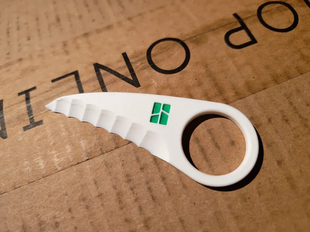 A white, serrated cutting tool with a circular handle rests on a piece of cardboard, featuring a small green logo on its side. The background shows the textured surface of the cardboard, with the words "TOP ONLY" partially visible.