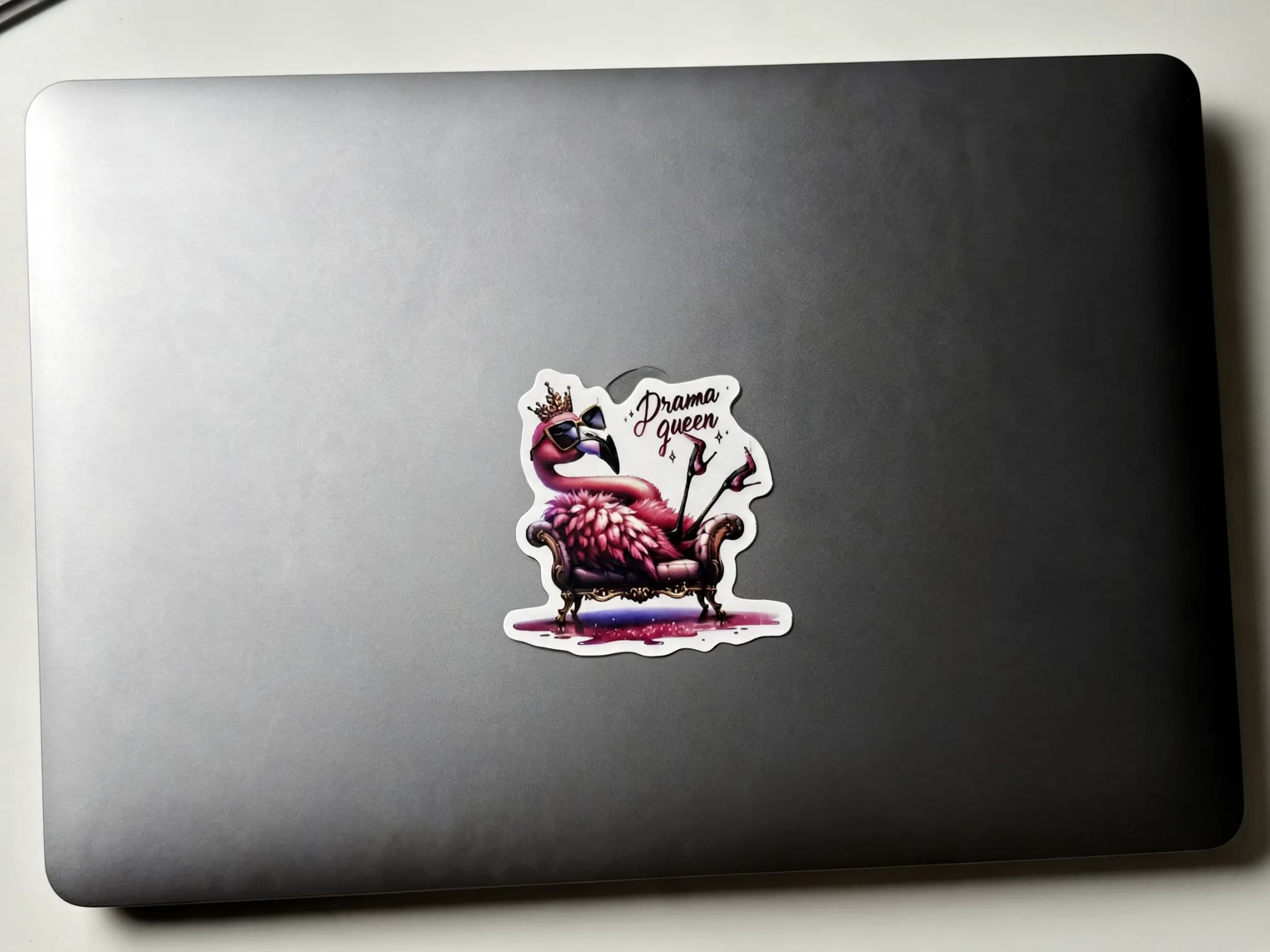 A colorful sticker of a flamingo wearing sunglasses and a crown, sitting on a chair with the text "Drama queen" above it, is placed on a dark laptop.