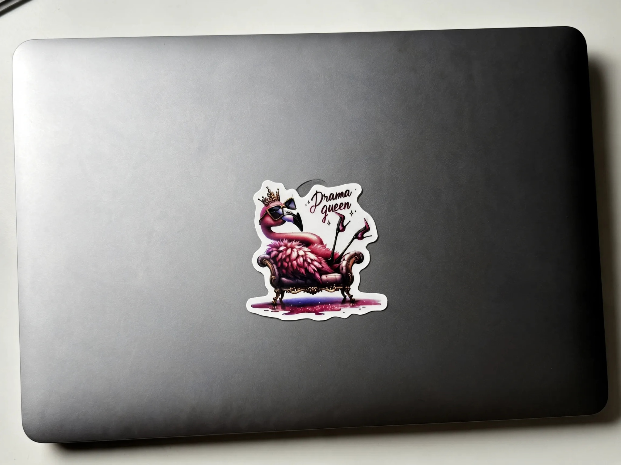 A colorful sticker of a flamingo wearing sunglasses and a crown, sitting on a chair with the text "Drama queen" above it, is placed on a dark laptop.