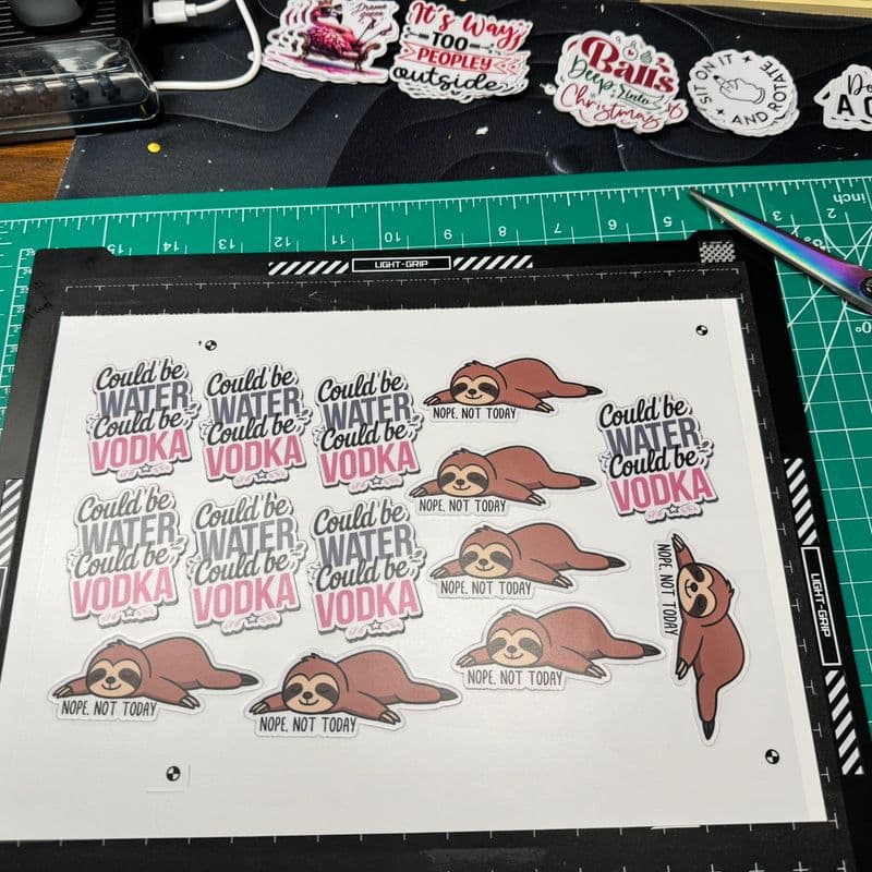 A cutting mat displays several colorful stickers with phrases like "Could be WATER, could be VODKA" and illustrations of sloths with the text "NOPE, NOT TODAY." Scissors are placed nearby.