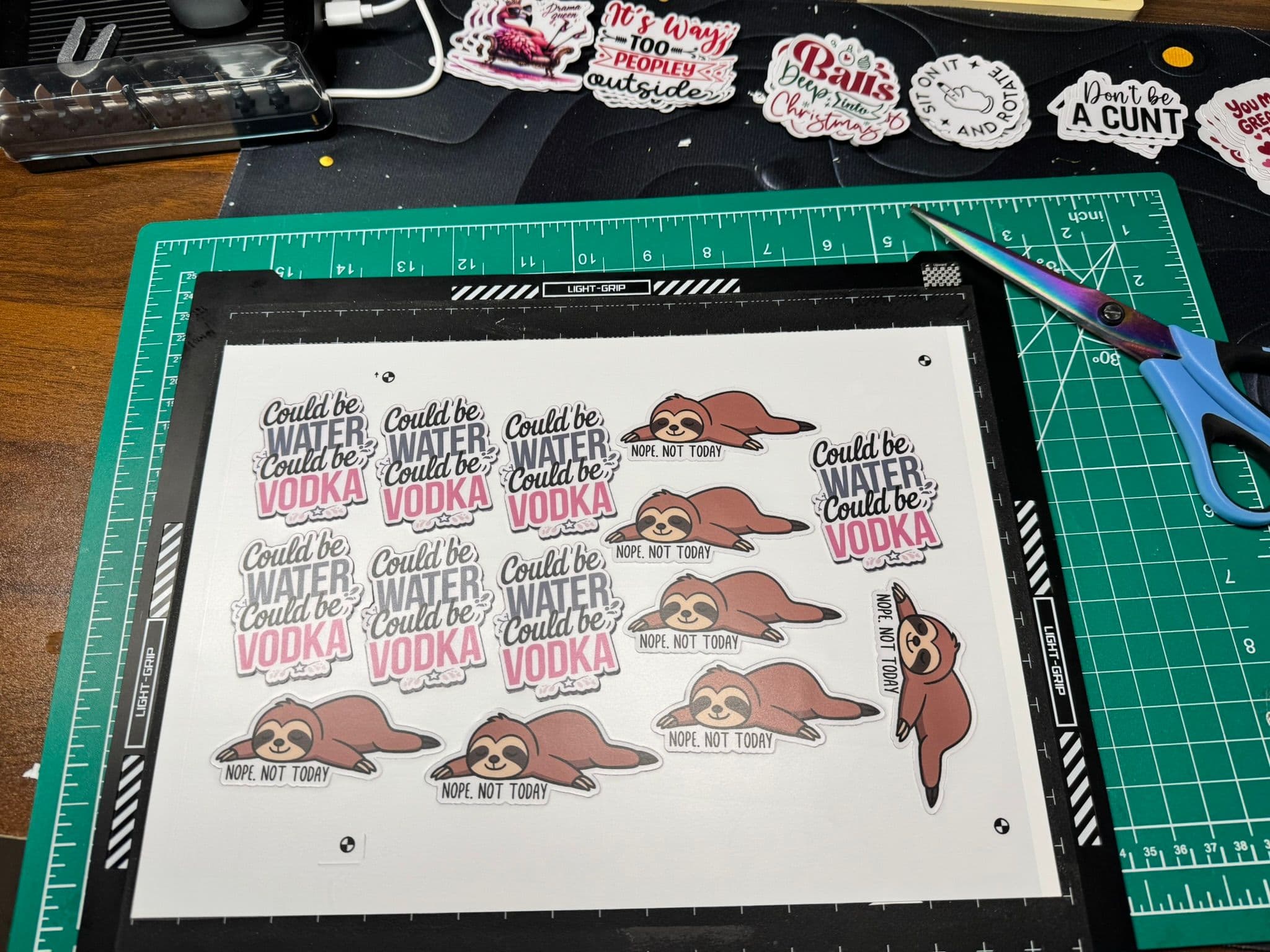 A cutting mat displays several colorful stickers with phrases like "Could be WATER, could be VODKA" and illustrations of sloths with the text "NOPE, NOT TODAY." Scissors are placed nearby.