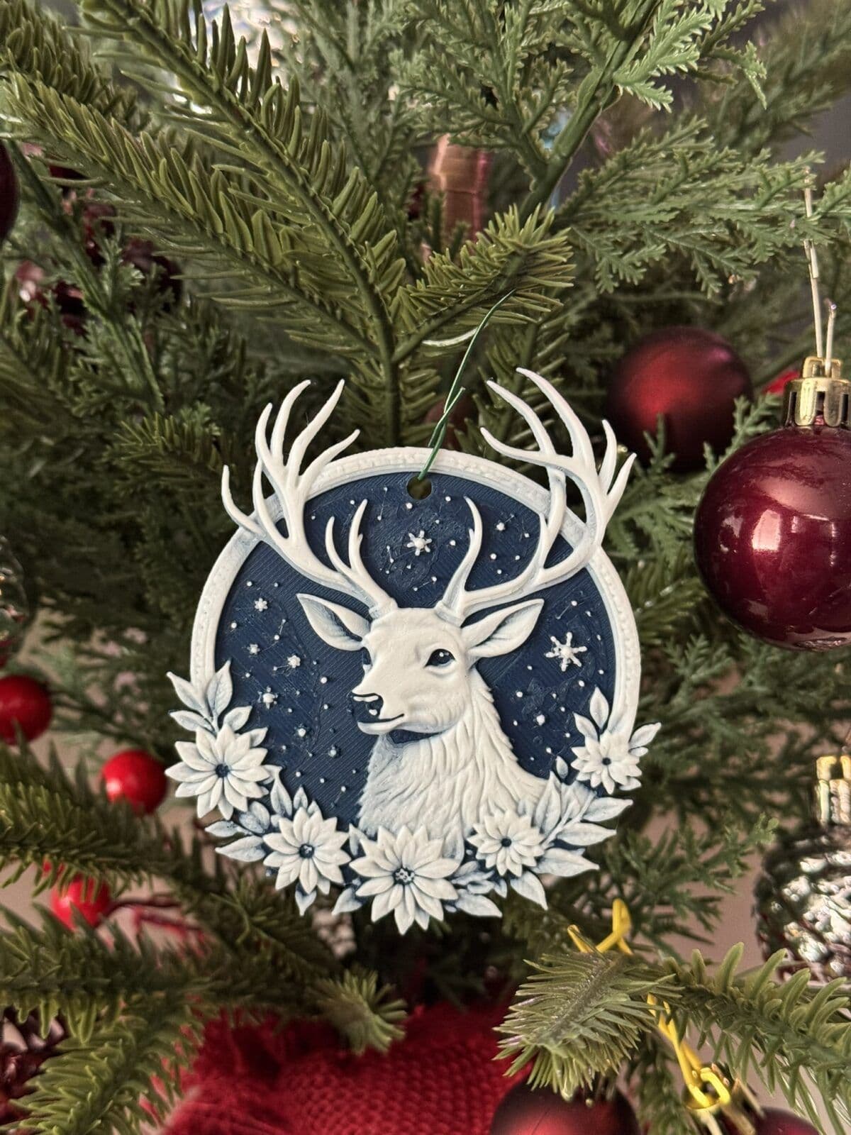 A decorative ornament featuring a white deer with prominent antlers, set against a dark blue circular background adorned with stars. The deer is surrounded by white flowers, hanging from a green branch of a Christmas tree, alongside red and silver ornaments.