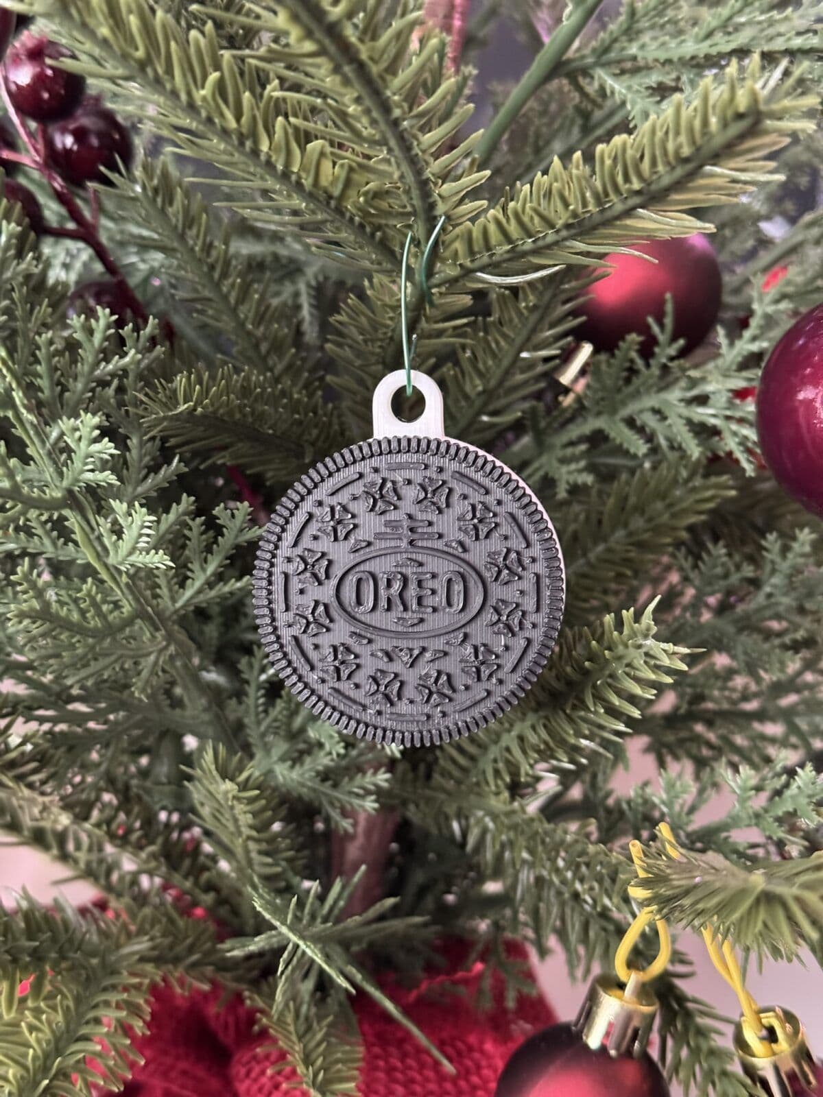 A black Oreo cookie ornament hangs from a green pine branch, surrounded by red ornaments and festive greenery. The ornament features detailed designs typical of an Oreo cookie, adding a playful touch to the holiday decor.
