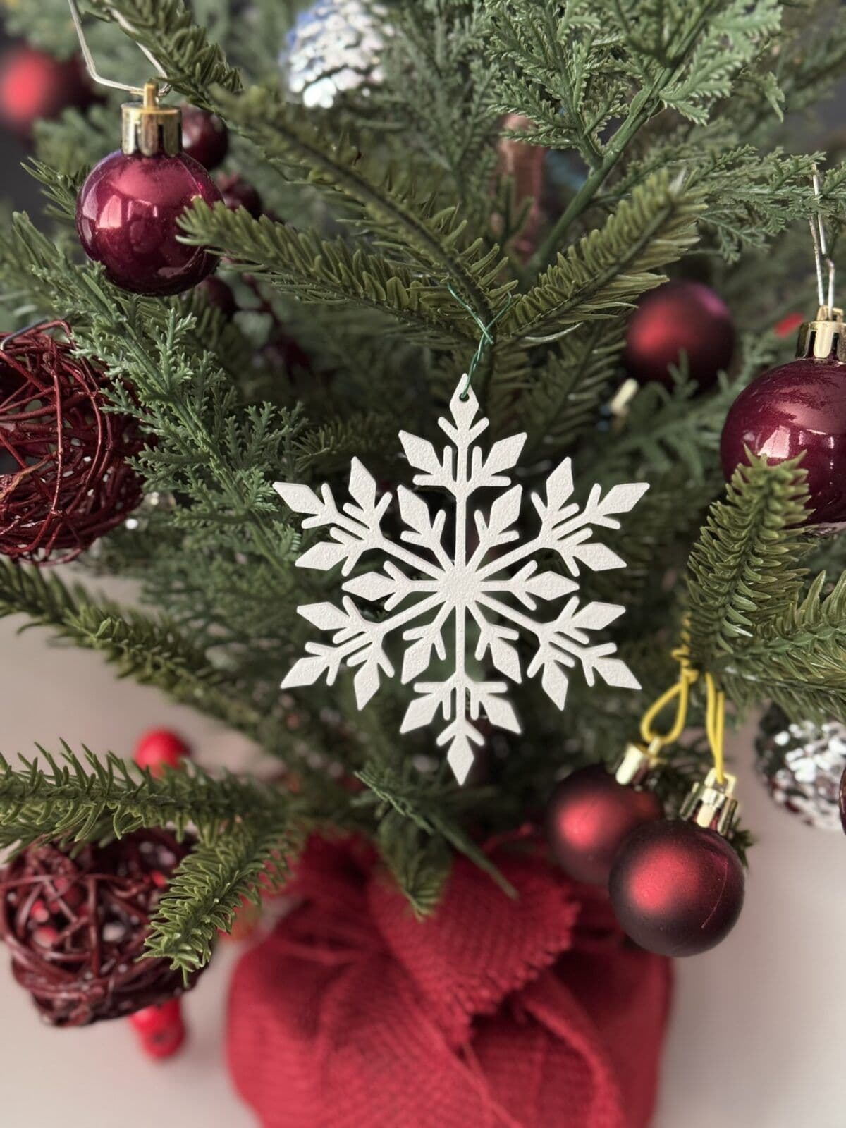 A small Christmas tree adorned with red and silver ornaments, including a prominent white snowflake decoration, is set against a neutral background. The tree is nestled in a red fabric base, enhancing the festive atmosphere.