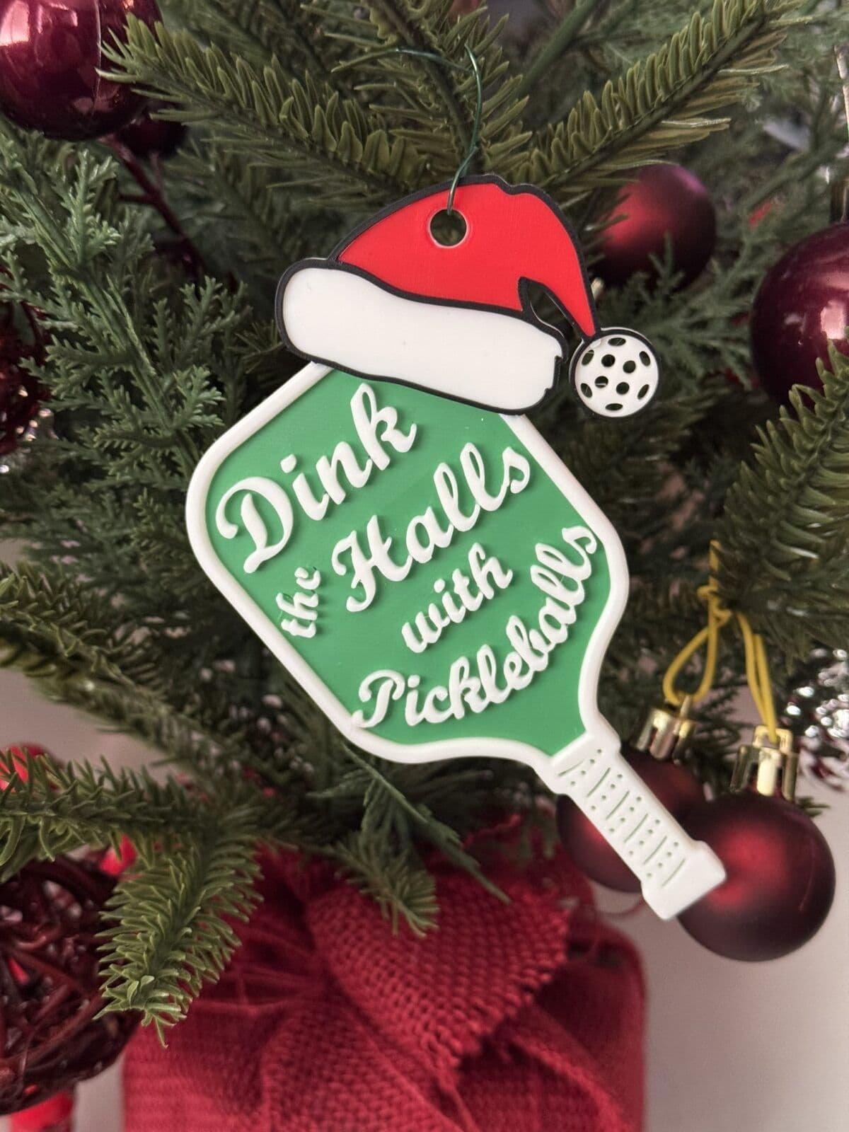 A festive ornament shaped like a pickleball paddle, featuring the phrase "Dink the Halls with Pickleballs" in playful white lettering. The ornament is adorned with a small Santa hat and hangs among green pine branches and red holiday decorations.