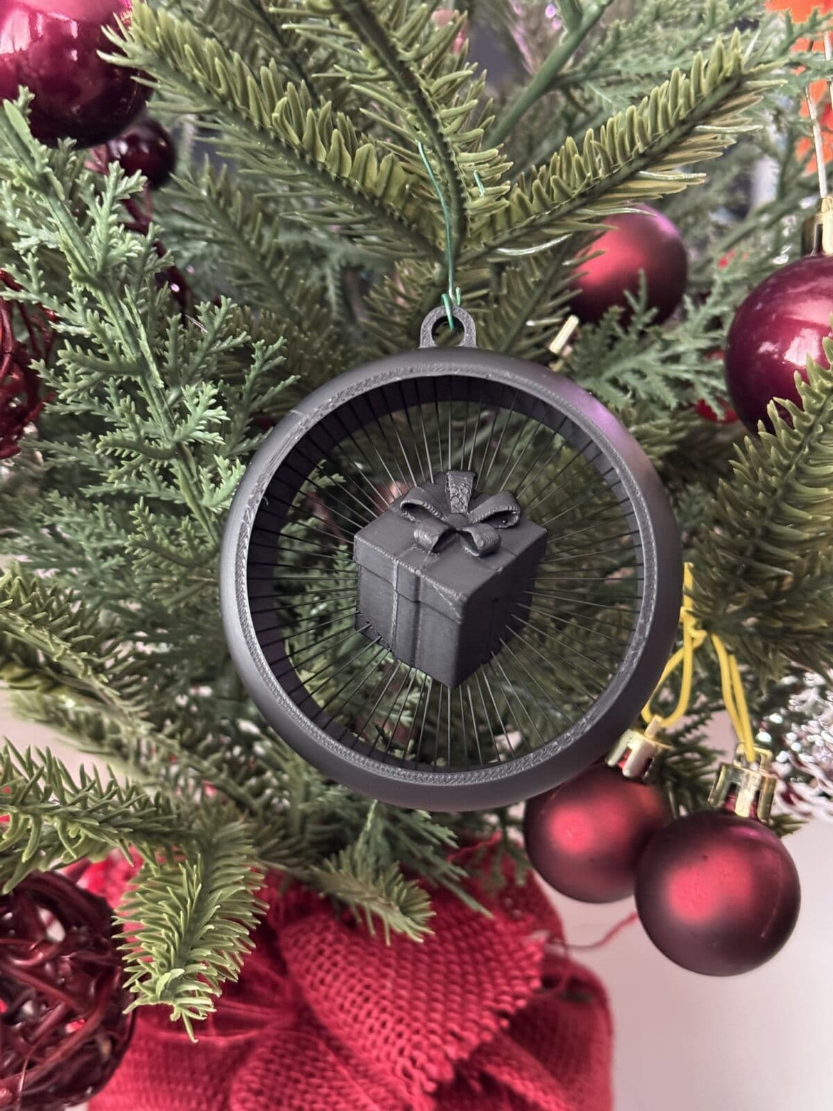 A black gift box with a bow is suspended inside a circular ornament, surrounded by green pine branches and red ornaments on a Christmas tree. The backdrop features a textured red fabric, enhancing the festive atmosphere.
