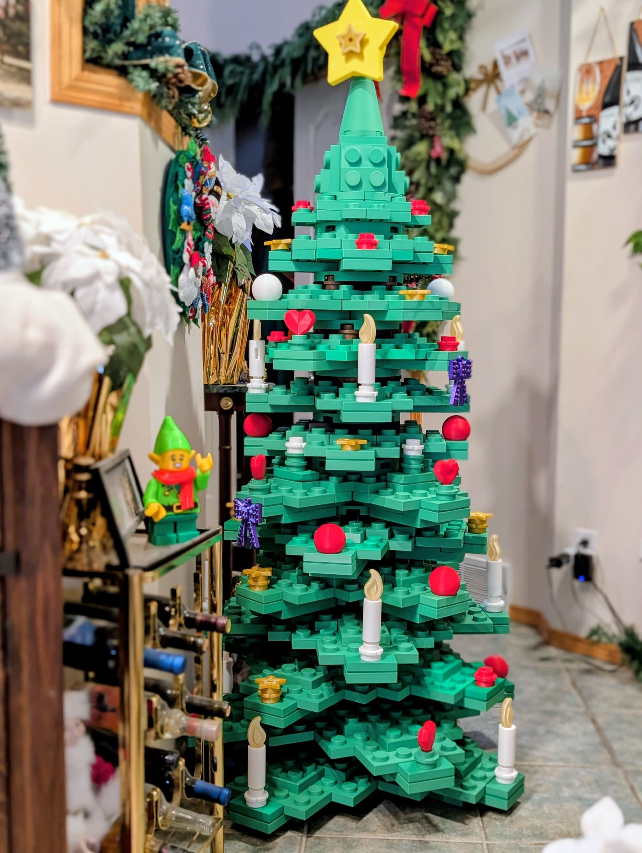 A colorful, Lego-style Christmas tree stands in a cozy indoor setting, adorned with red and white ornaments, candles, and a star on top. In the foreground, a small Lego elf figure with a green hat and red scarf is positioned next to a wine rack filled with bottles.