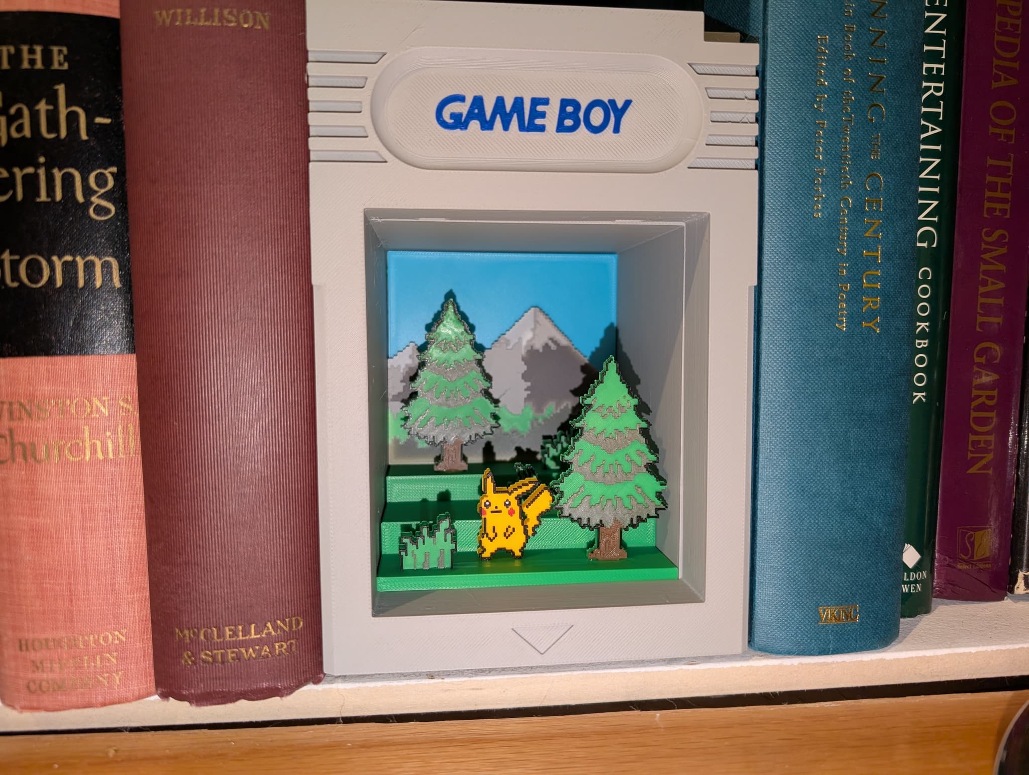 A colorful diorama featuring Pikachu standing in a green landscape with trees and mountains in the background, framed within a retro Game Boy console. The scene is displayed on a shelf lined with various books.