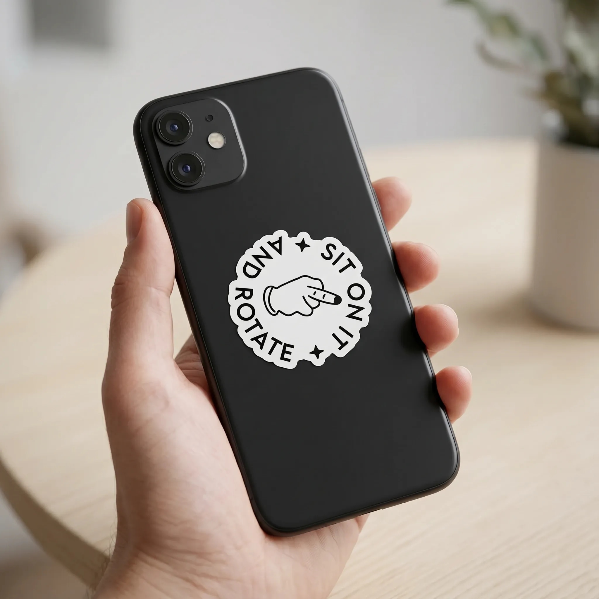A black smartphone is held in a hand, featuring a circular sticker with a pointing hand and the text "SIT ON IT AND ROTATE."