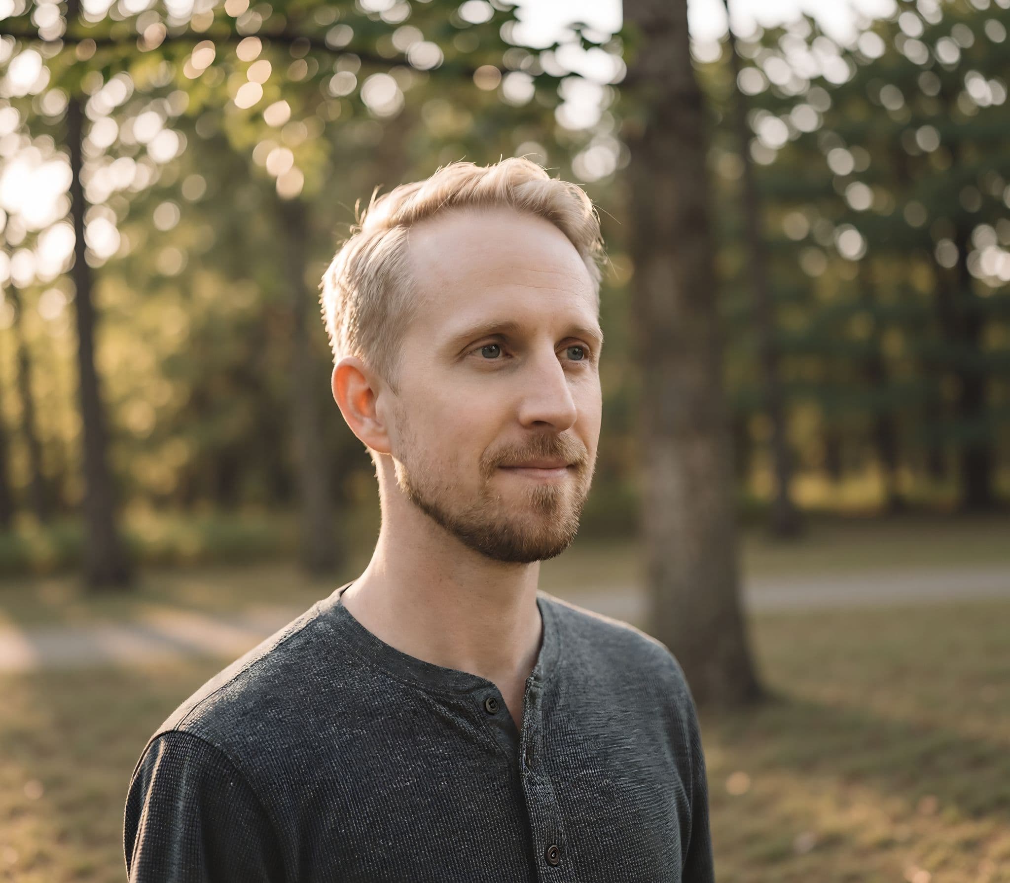 A man with short blonde hair and a light beard stands outdoors, looking thoughtfully into the distance. The background features softly blurred trees and warm sunlight filtering through the leaves, creating a serene atmosphere.