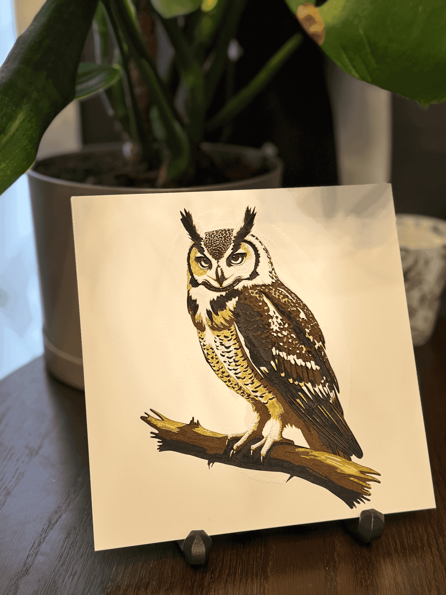 A detailed 3d printed filament art featuring an owl perched on a branch, showcasing its distinctive features such as large eyes and tufted ears. The artwork is displayed on a stand against a backdrop of green plants and soft lighting.