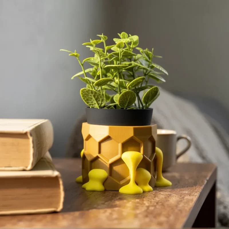 A vibrant green plant sits in a yellow, honeycomb-patterned pot with playful yellow supports, placed on a wooden table alongside two stacked books and a beige mug. In the background, a soft, neutral-toned couch is partially visible, adding a cozy ambiance to the scene.