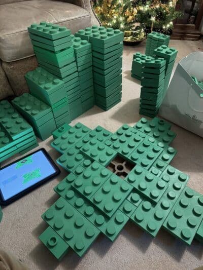 A collection of green building blocks is spread out on a carpeted floor, with several stacks neatly arranged nearby. An electronic device displaying a building plan is positioned next to the blocks, indicating a construction project in progress.