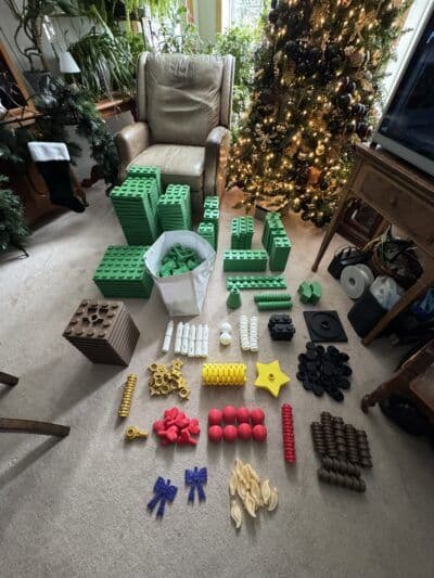 A variety of colorful building blocks and construction pieces are arranged on a carpeted floor, with green blocks stacked in groups and other shapes in various colors, including red, yellow, and black, spread out around them. In the background, a decorated Christmas tree adds a festive touch to the cozy living room setting.