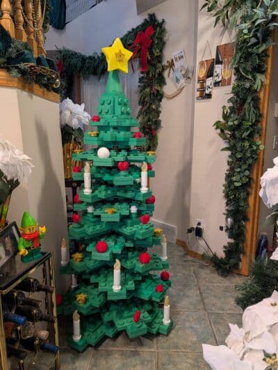 A colorful, large Christmas tree made of green building blocks stands in a festive entryway, adorned with red ornaments, white candles, and topped with a yellow star. Surrounding decorations include garlands, white flowers, and holiday-themed wall art, creating a cheerful atmosphere.