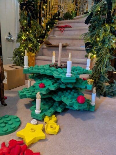 A colorful, block-style Christmas tree made from green building blocks stands on a carpeted floor, adorned with white candle replicas and red ornaments. In the background, festive decorations and a staircase are visible, enhancing the holiday atmosphere.