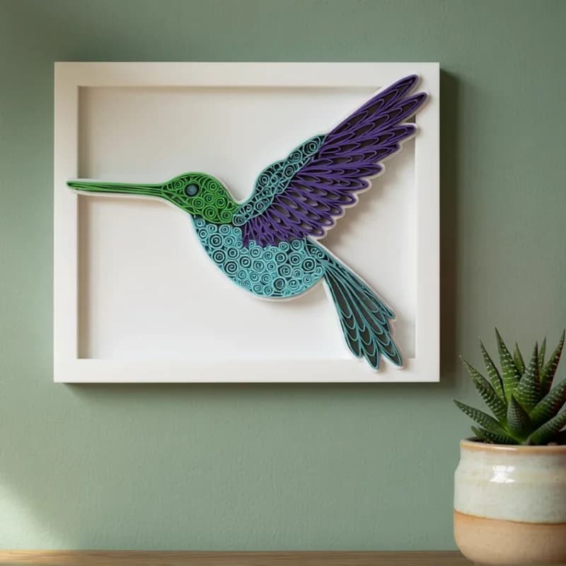 A vibrant, three-dimensional paper art piece of a hummingbird is displayed in a white frame against a soft green wall. The bird features intricate patterns in shades of blue, green, and purple, with a small potted succulent and colorful books visible on a wooden surface nearby.
